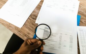 Mão segurando lupa sobre extratos e demonstrativos financeiros em uma mesa, representando a revisão fiscal para recuperação de créditos tributários